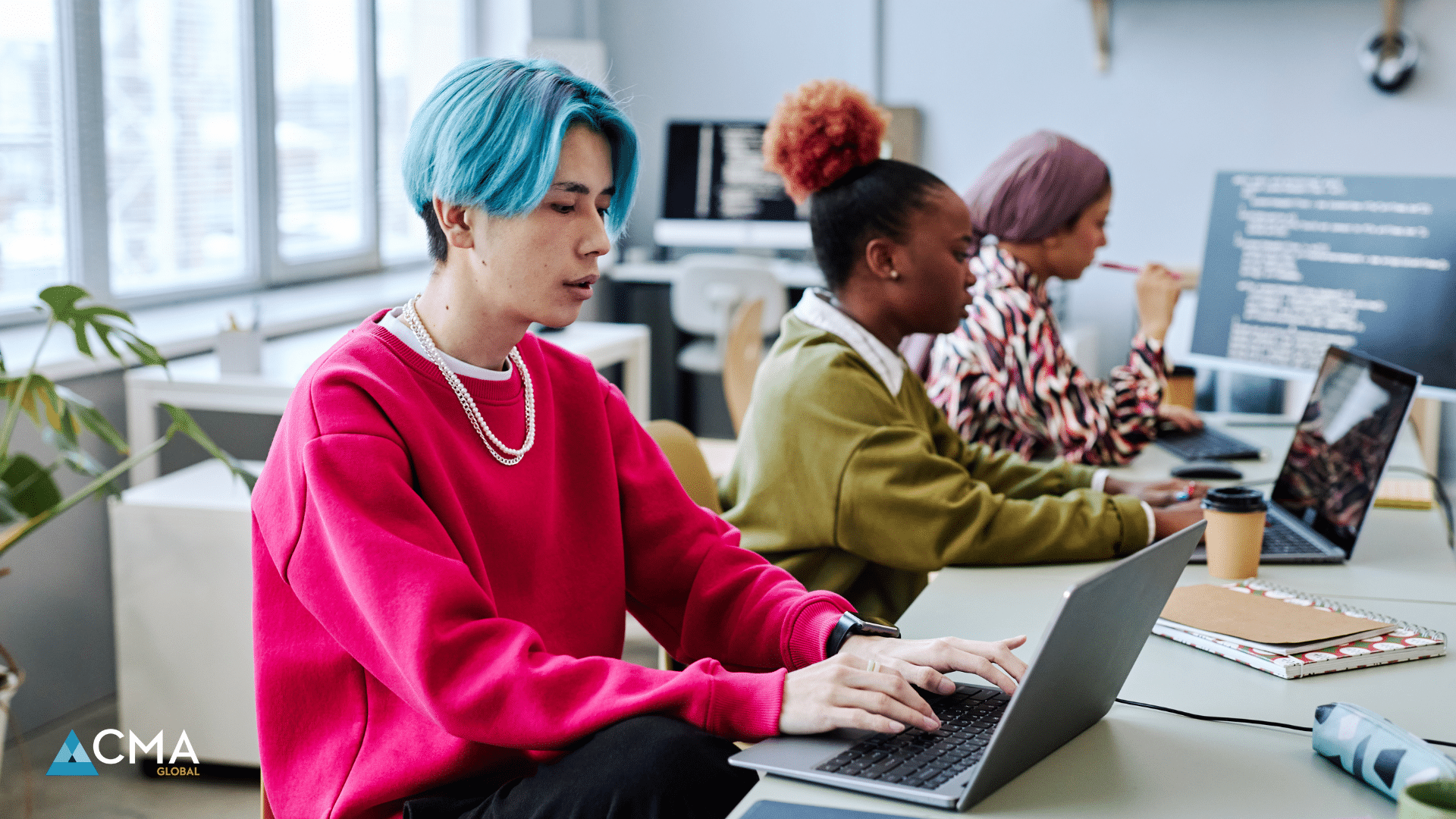 Three Gen-Z professionals work at laptops in a modern office space. In the foreground, a person with bright blue hair wears a vibrant red sweatshirt and silver chain necklace. Behind them, two colleagues with colorful hair - one with a red updo and another with purple hair - focus on their work, representing the diverse, contemporary workplace culture of younger generations.